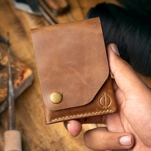 May include: A tan brown leather wallet featuring a fold-over flap secured by a single brass-coloured metal snap button. The wallet has a smooth, matte texture and includes a small circular logo embossed on the bottom right corner of the front panel.
