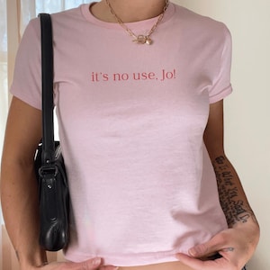 May include: A pink t-shirt with the text "it's no use, Jo!" printed in red on the front. The shirt is being worn by a person with a black leather bag slung over their shoulder.
