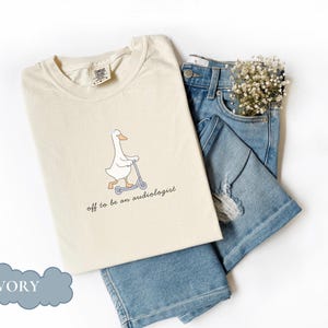 May include: Ivory-colored t-shirt with a cartoon goose riding a scooter, the text reads "off to be an audiologist." The shirt is paired with blue denim shorts and a sprig of white flowers.