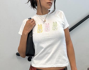May include: White t-shirt with a graphic of three pastel colored bunnies holding flowers. The bunnies are yellow, pink, and green.