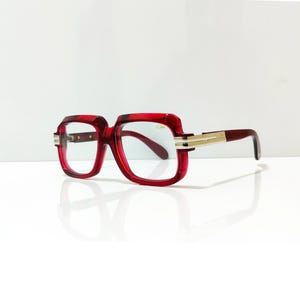 以下が含まれることがあります： 四角い形の赤いフレームの眼鏡。テンプルとヒンジには金色のアクセントがあります。レンズは透明で、フレームは深い赤色です。白い背景に配置されています。