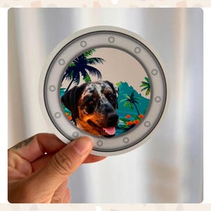 A 4x4 inch round magnet being held up in between a woman's fingers. The magnet is a ship's porthole with a tropical background and has a custom dog portrait inside.