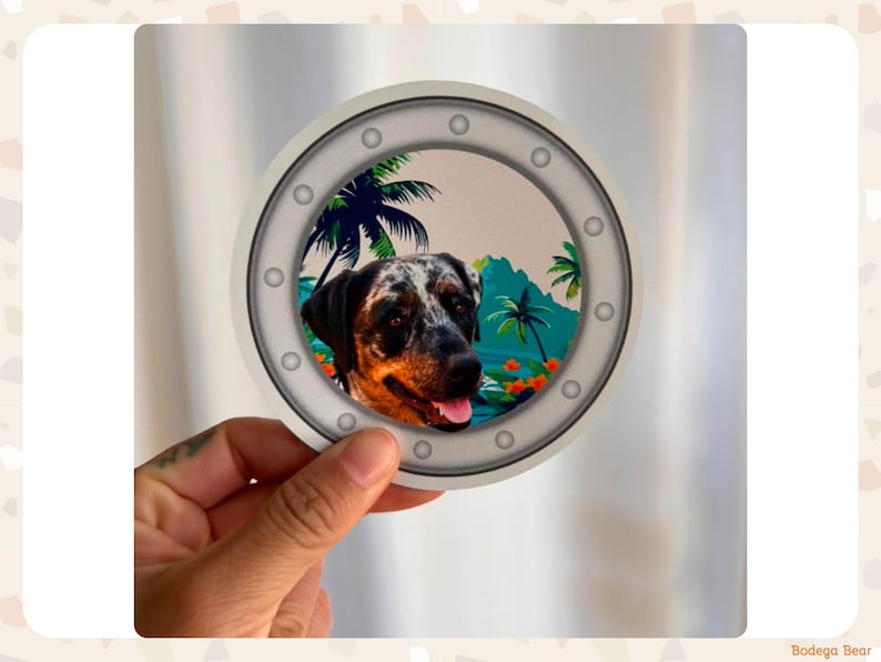 A 4x4 inch round magnet being held up in between a woman's fingers. The magnet is a ship's porthole with a tropical background and has a custom dog portrait inside.
