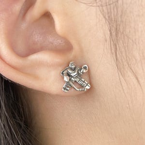 May include: A silver hockey player stud earring. The earring features a hockey player in a playing position, complete with a hockey stick and protective equipment. The earring is small and detailed, suitable for everyday wear.
