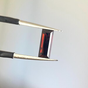 May include: A rectangular, deep red gemstone is held by a pair of tweezers. The gem has a polished, faceted surface, reflecting light. The tweezers have black handles and silver tips. The background is a soft, neutral gray.