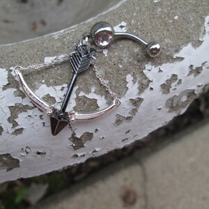 May include: A silver body piercing with a black arrow and bow charm. The arrow is pointing upwards and the bow is curved. The piercing is on a white background.
