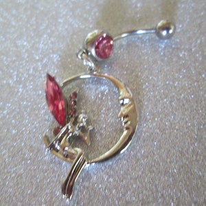 May include: A silver belly button ring with a pink crystal on the top and a dangling charm featuring a crescent moon with a face and a fairy with pink crystals.