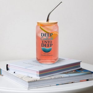 May include: A clear glass can-shaped tumbler filled with a pink beverage, fruit slices, and a stainless steel straw. The tumbler has a wooden lid and features the text "DEEP Calleth UNTO DEEP" with a wave graphic.