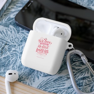 May include: White AirPods case with a silver carabiner clip, open to reveal white earbuds. The case features the text "We love because He first loved us" in pink script. The case is on a blue and white patterned surface.