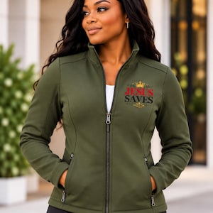 May include: An olive green zip-up jacket featuring a high collar and two zippered side pockets. The left chest displays a graphic with a gold crown above the red text "JESUS" and black text "SAVES" with a small gold cross icon underneath.