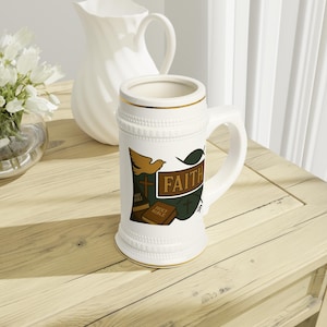 May include: White ceramic beer stein with a gold rim and handle. The stein features a design with the word "FAITH", a dove, a cross, and a Bible. The stein is on a light wood surface.