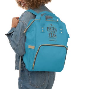 Peut inclure: Un sac à dos bleu vif doté d'une poignée supérieure et d'une poche avant zippée. Le sac affiche le texte FAITH OVER FEAR 2 TIMOTHY 1:7 et HolyWordThreads en lettres marron foncé avec un symbole de croix au-dessus des mots sur le panneau avant.