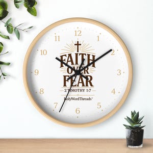 May include: A circular wall clock with a light wood surround. The white clock face has black hands and numerals. The centre displays the brown text "FAITH OVER FEAR", a cross, and the scripture reference 2 Timothy 1:7.