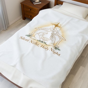 Könnte beinhalten: Weiße Decke mit goldfarbener Illustration einer Krippenszene mit Stern und dem Text "Silent Night, Holy Night". Die Decke liegt auf einem Bett mit Holzrahmen. Im Hintergrund ist ein Nachttisch zu sehen.