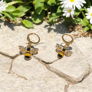 May include: A pair of gold-tone bee earrings. Each earring features a yellow and black bee pendant with crystal-like accents on the wings. The earrings are set against a stone surface with greenery and white flowers in the background.