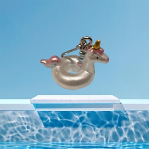 May include: A small unicorn float charm with a pearlescent finish, featuring a pink mane and tail, and a gold horn. The charm is suspended by a silver-coloured metal ring. The background is a gradient of blue.