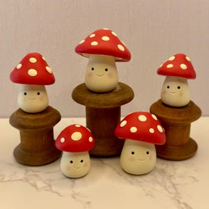 Puede incluir: Cinco figuritas de setas de arcilla hechas a mano con sombreros pintados de rojo y blanco y caras sonrientes. Las setas se muestran en carretes de madera.