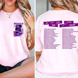 May include: Light pink t-shirt featuring a purple graphic on the front with the text "World Tour 2026" and faces. The back of the shirt lists tour dates and locations in purple text, including cities like Toronto, Chicago, and Los Angeles.