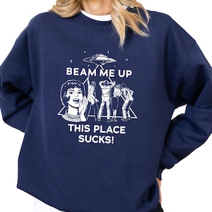 May include: Navy blue sweatshirt with a white graphic that reads "BEAM ME UP THIS PLACE SUCKS!" The graphic depicts a retro woman, a UFO, and figures being beamed up. The sweatshirt has a crew neck and long sleeves.