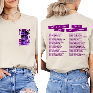 May include: A beige t-shirt featuring a purple graphic with the text "World Tour 2026" and band member images. The back of the shirt lists tour dates and locations. The shirt is paired with blue jeans.