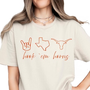 May include: Cream-colored t-shirt with the phrase "hook 'em horns" in a burnt orange color. The design includes a hand gesture, a Texas outline, and a longhorn silhouette.