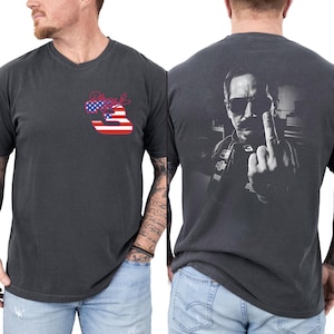 May include: A charcoal gray short-sleeve t-shirt featuring a small graphic on the front chest with the number 3 in a red, white, and blue American flag pattern. The back displays a large black and white portrait of a person wearing sunglasses.