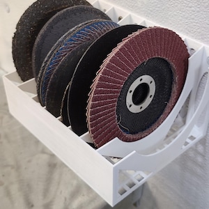 May include: A white plastic wall-mounted storage rack with six slots for holding sanding discs. The rack is holding five sanding discs, four black and one red.