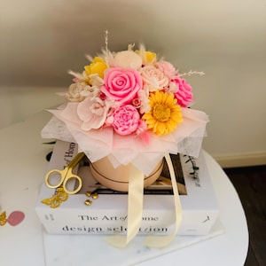 Può includere: Una composizione floreale in una scatola rotonda rosa chiaro con un nastro color crema. Il bouquet presenta rose rosa, girasoli gialli e altri fiori colorati. La scatola è appoggiata su una pila di libri con forbici dorate ed elementi decorativi.