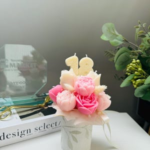 May include: A floral arrangement of pink and cream-colored rose-shaped candles, with the number "18" in candles. The arrangement is in a small, decorative, cylindrical container with a leaf pattern. A ribbon is tied around the container.