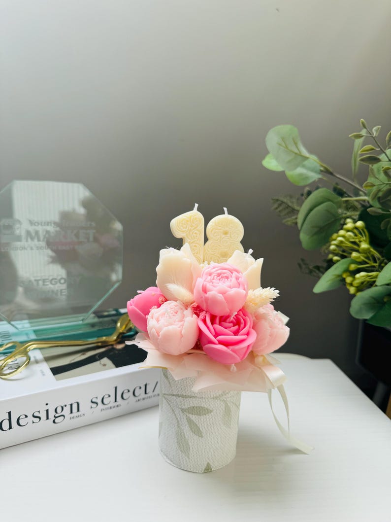 May include: A floral arrangement of pink and cream-colored rose-shaped candles, with the number "18" in candles. The arrangement is in a small, decorative, cylindrical container with a leaf pattern. A ribbon is tied around the container.