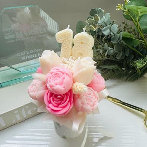 May include: A floral arrangement featuring pink and cream-colored peony-shaped candles, with a cream-colored number 18 candle. The arrangement is in a small, decorative pot with a white ribbon. Greenery and a gold-colored pair of scissors are in the background.