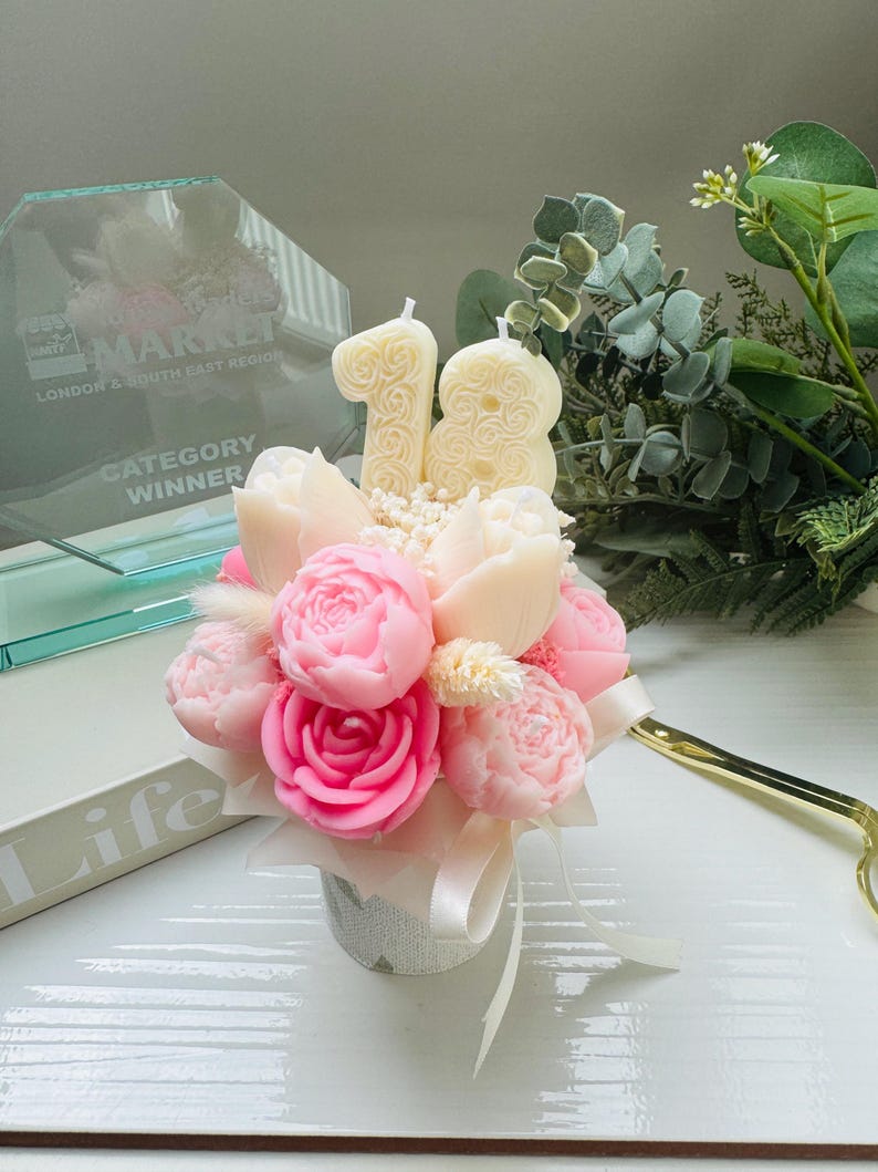 May include: A floral arrangement featuring pink and cream-colored peony-shaped candles, with a cream-colored number 18 candle. The arrangement is in a small, decorative pot with a white ribbon. Greenery and a gold-colored pair of scissors are in the background.