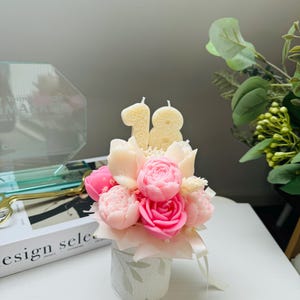 May include: A floral arrangement of pink and white candles shaped like roses and other flowers, with the number "18" on top. The arrangement is in a small, decorative, white and green floral patterned container. Ribbons are tied around the container.