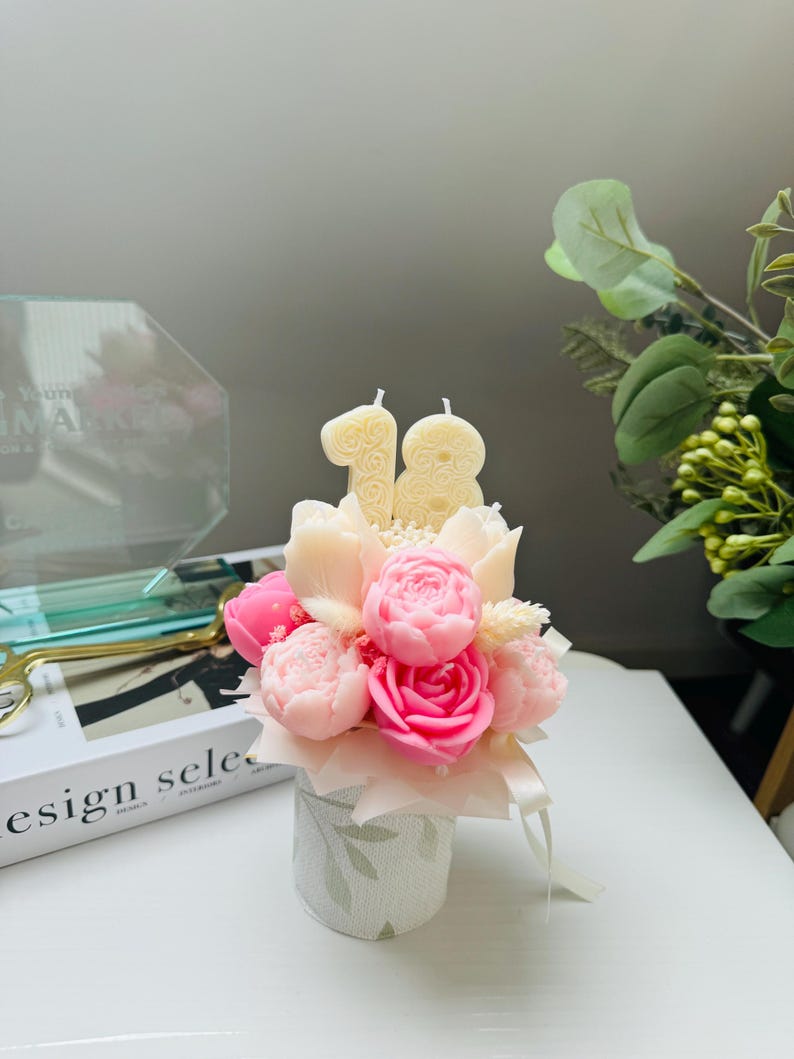 May include: A floral arrangement of pink and white candles shaped like roses and other flowers, with the number "18" on top. The arrangement is in a small, decorative, white and green floral patterned container. Ribbons are tied around the container.