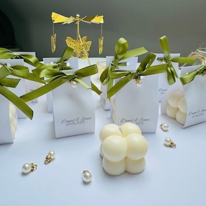 May include: White gift boxes with green satin ribbon and pearl embellishments. A gold butterfly mobile and bubble candles are visible. The boxes are labelled with names and a date.