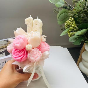 May include: A floral arrangement featuring pink and white flower-shaped candles, including roses and peonies, with the number "18" in candles. The bouquet is tied with a white ribbon and presented in a small, textured vase.