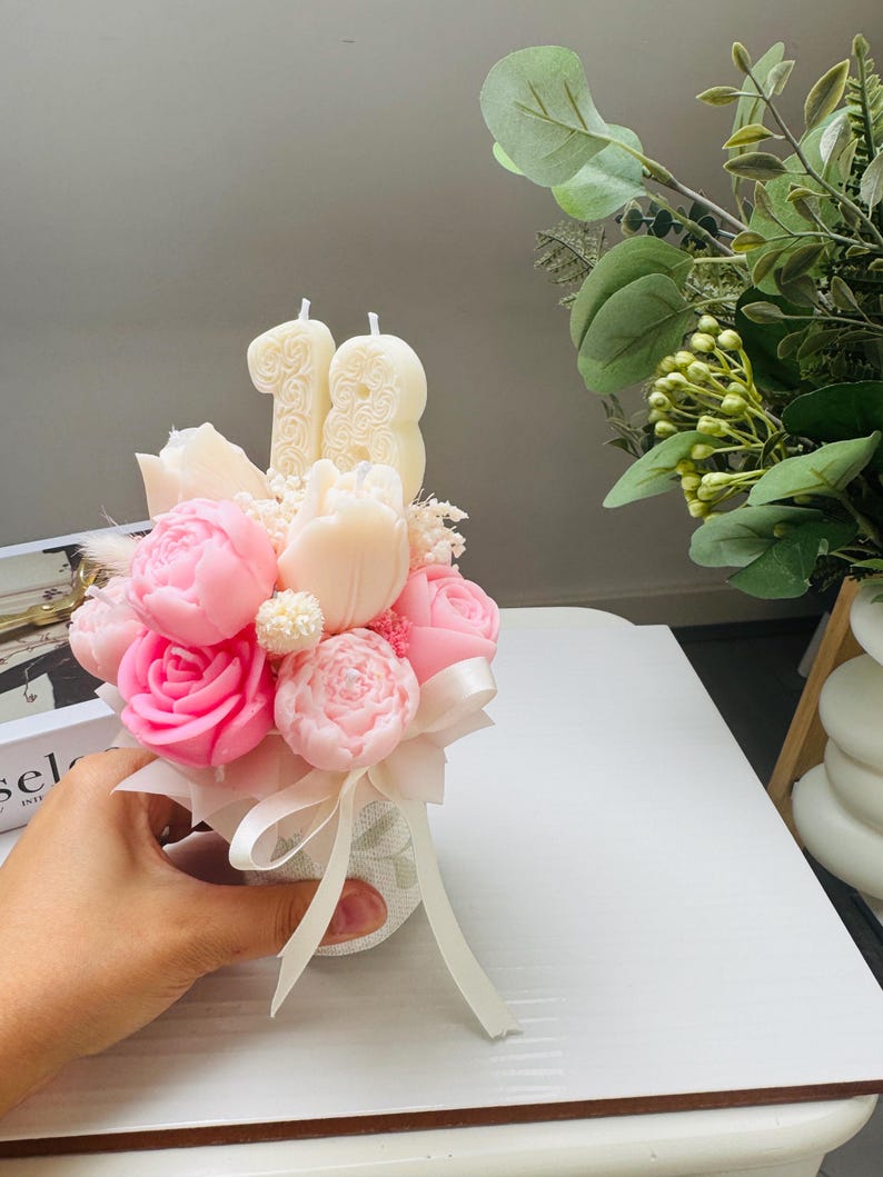 May include: A floral arrangement featuring pink and white flower-shaped candles, including roses and peonies, with the number "18" in candles. The bouquet is tied with a white ribbon and presented in a small, textured vase.