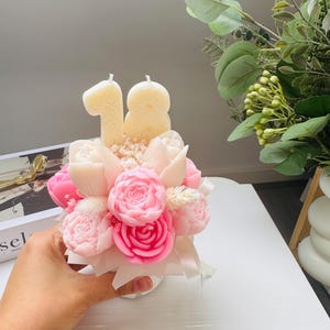 May include: A floral arrangement candle with pink and white roses, tulips, and small white flowers. Two cream-colored candles shaped as the number 18 are placed above the flowers. The arrangement is held in a small white vase and tied with a white ribbon.