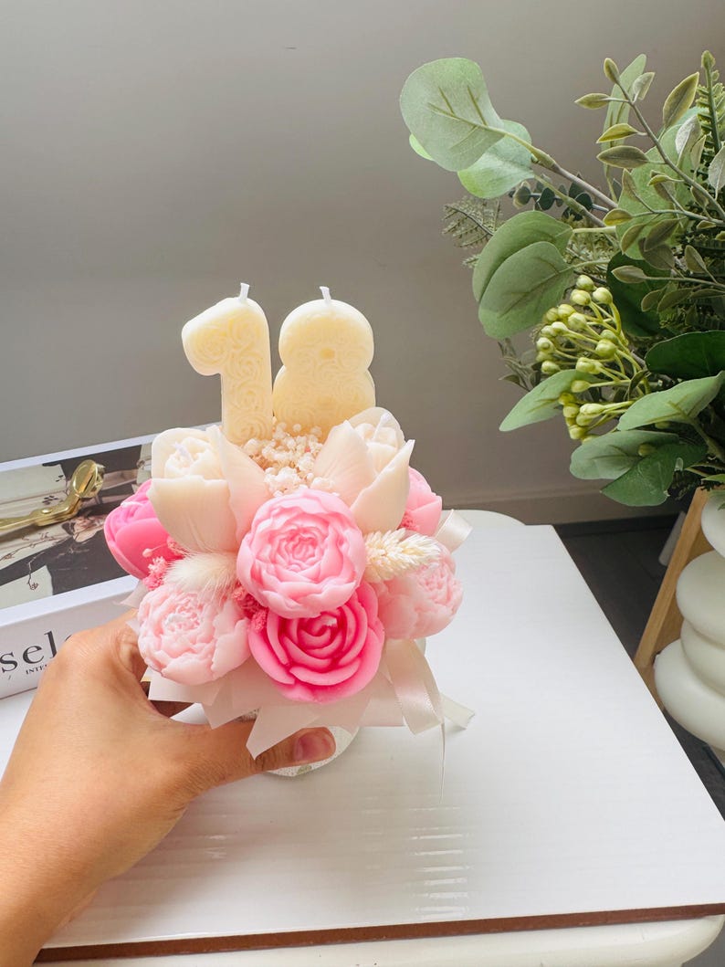May include: A floral arrangement candle with pink and white roses, tulips, and small white flowers. Two cream-colored candles shaped as the number 18 are placed above the flowers. The arrangement is held in a small white vase and tied with a white ribbon.