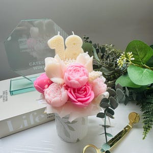 May include: A floral arrangement of pink and cream-colored peony-shaped candles in a small, patterned vase. Two cream-colored number candles, 1 and 8, are placed above the flowers. A glass award and gold-colored scissors are also visible.