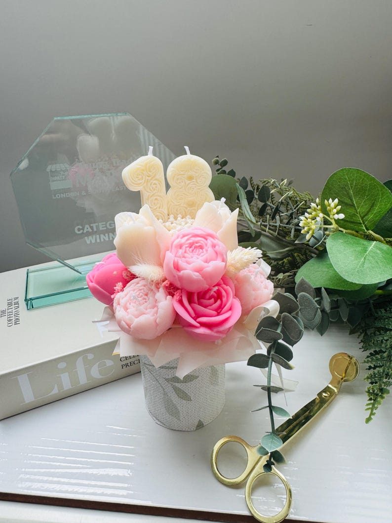 May include: A floral arrangement of pink and cream-colored peony-shaped candles in a small, patterned vase. Two cream-colored number candles, 1 and 8, are placed above the flowers. A glass award and gold-colored scissors are also visible.