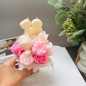 May include: A floral arrangement of pink and white rose-shaped candles, with the number "18" in candle form on top. The arrangement is in a small white box, held by a hand. Greenery is visible in the background.