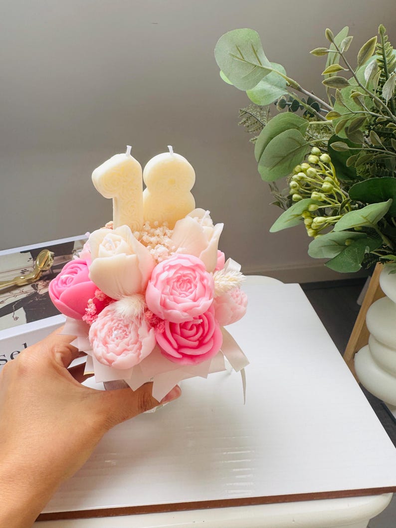 May include: A floral arrangement of pink and white rose-shaped candles, with the number "18" in candle form on top. The arrangement is in a small white box, held by a hand. Greenery is visible in the background.