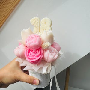 May include: A floral arrangement of pink peony and rose-shaped candles, with two number 18 candles, in a small, decorative container. The arrangement is tied with a white ribbon.