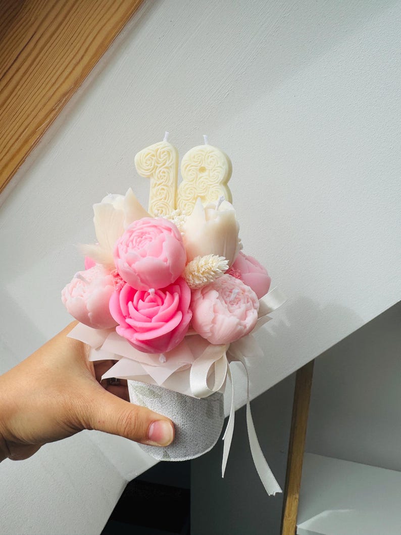 May include: A floral arrangement of pink peony and rose-shaped candles, with two number 18 candles, in a small, decorative container. The arrangement is tied with a white ribbon.