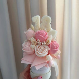 May include: A floral arrangement featuring pink and white roses, tulips, and small white flowers. The bouquet is accented with a pink bow and the number "18" in white. The arrangement is presented in a white container.