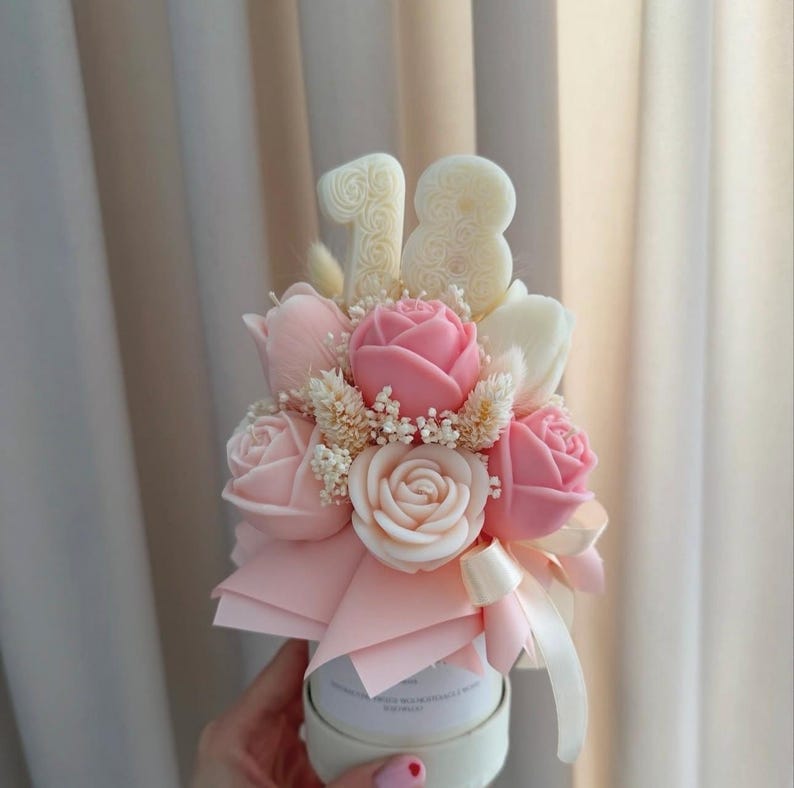 May include: A floral arrangement featuring pink and white roses, tulips, and small white flowers. The bouquet is accented with a pink bow and the number "18" in white. The arrangement is presented in a white container.