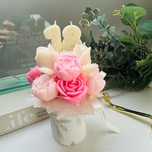 May include: A floral arrangement featuring pink and white peony and rose-shaped candles, with two candles shaped as the number 18. The arrangement is in a small, patterned container with a ribbon. Greenery and a gold-colored object are in the background.