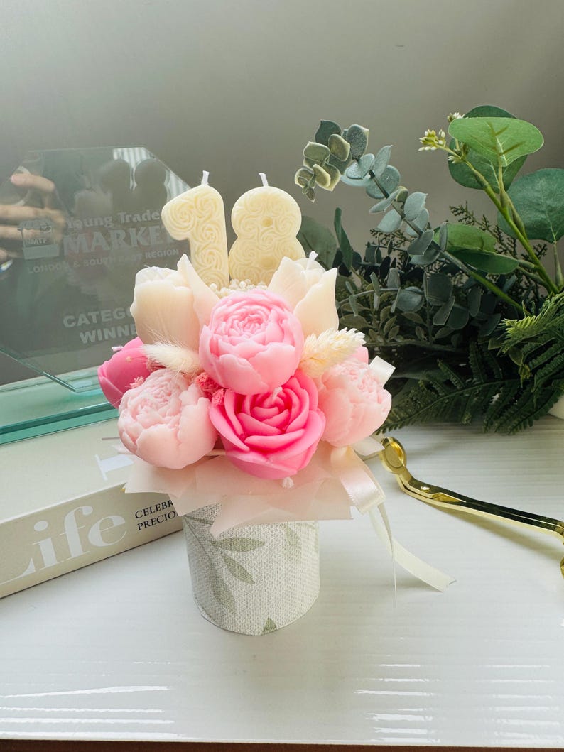 May include: A floral arrangement featuring pink and white peony and rose-shaped candles, with two candles shaped as the number 18. The arrangement is in a small, patterned container with a ribbon. Greenery and a gold-colored object are in the background.