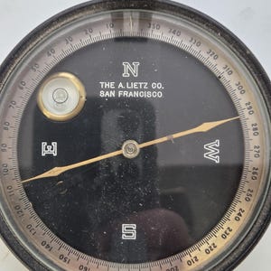 May include: A vintage black compass with a clear glass cover. The compass face has degree markings, cardinal directions, and the text "THE A. LIETZ CO. SAN FRANCISCO." A bubble level is visible. The compass needle points east.